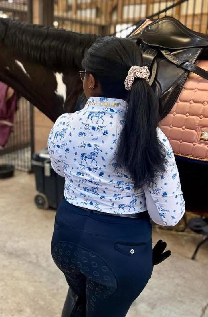 A woman in equestrian apparel stands in a horse stable next to a dark brown and white horse with a saddle on its back. The woman is wearing a white long-sleeve shirt with blue horse motifs and dark navy breeches.